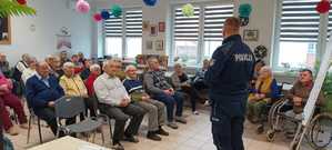 Policjant na spotkaniu z seniorami stojąc przy tablicy ostrzega seniorów przed oszustami.