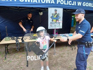 Dziecko w policyjnym kasku na głowie i trzymające policyjną tarczę, obok stoi policjant.