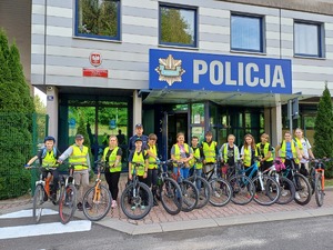 Policjant wraz z dziećmi trzymającymi rowery stoi przed wejściem do komendy policji.