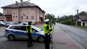 Policjant z miernikiem prędkości kontroluje prędkość jadących pojazdów na drodze. Obok stoi drugi policjant i radiowóz.