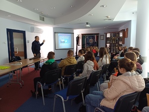 Grupa młodzieży słuch prelekcji policjanta na auli, na ekranie wyświetlany jest slajd.