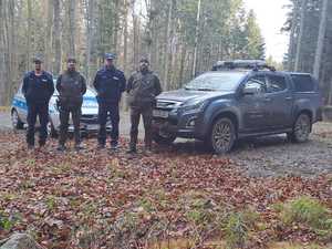 Na polanie stoi samochód policyjny i straży leśnej, a obok stoi dwóch policjantów i dwóch strażników leśnych.