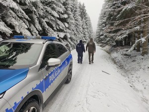 Radiowóz policyjny stoi na ośnieżonej drodze, za samochodem stoją policjant i strażnik leśny.