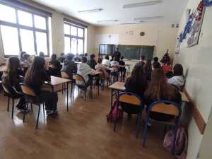 W sali szkolnej dzieci siedzą przy ławkach, a policjant przy tablicy prowadzi prelekcje.