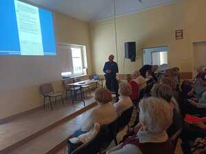 Policjant stoi i prowadzi prelekcje do siedzących na sali seniorów.