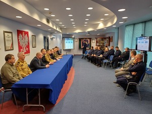 Przy stole siedzą przedstawiciele służb i instytucji powiatu gorlickiego. Na przeciw siedzi kadra kierownicza komendy policji w Gorlicach. Wszyscy patrzą na wyświetlaną prezentację.