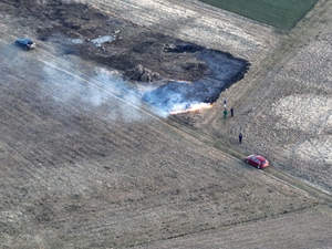 Widoczny z góry  wypalony obszar trawy. Obok stojące cztery osoby oraz dwa pojazdy.