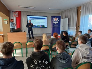 Policjant stojąc mówi do młodzieży. W tle na ekranie prezentacja dotycząca służby w policji.