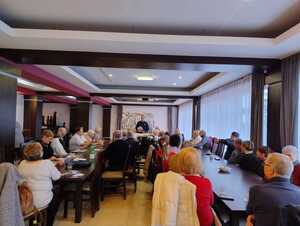 Seniorzy siedzą przy stołach i słuchają prelekcji policjanta.