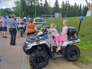Dwoje dzieci siedzi na quadzie, z tyłu stoi samochód i motocykl policyjny.