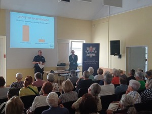 Siedzący na sali seniorzy słuchają wypowiedzi powiatowego rzecznika praw konsumenta.