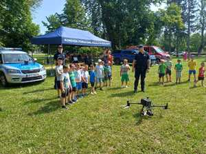 Dzieci razem z policjantem stoją koło policyjnego namiotu i obserwują start drona.