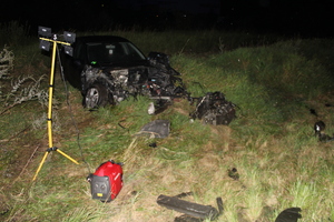 Na łące stoi rozbity samochód, a obok leżą różne elementy pojazdu.