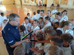 Policjant na sali gimnastycznej rozdaje odblaski dzieciom ubranym w kimona.