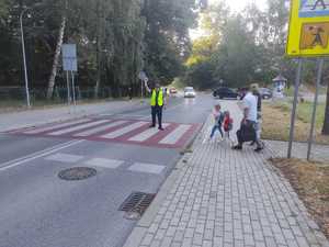 Policjant stoi na przejściu dla pieszych i daje kierującym sygnał lizakiem aby się zatrzymali bo przez przejście przechodzą dzieci.
