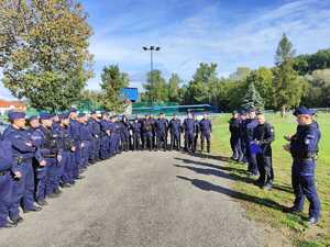 Obok stadionu sportowego stoi pododdział policjantów przed ćwiczeniami.