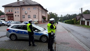 Policjanci stoją koło radiowozu i kontrolują prędkość jadących drogą pojazdów.
