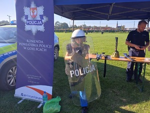 Obok policyjnego namiotu stoi chłopiec ubrany w policyjny strój ochronny z tarczą.