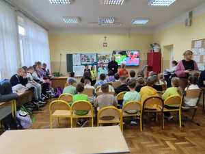 Uczniowie się siedzą na krzesłach w klasie i słuchają prelekcji policjantki.