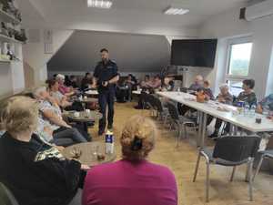 Policjant rozdaje uczestnikom spotkania, którzy siedzą przy stołach elementy odblaskowe.