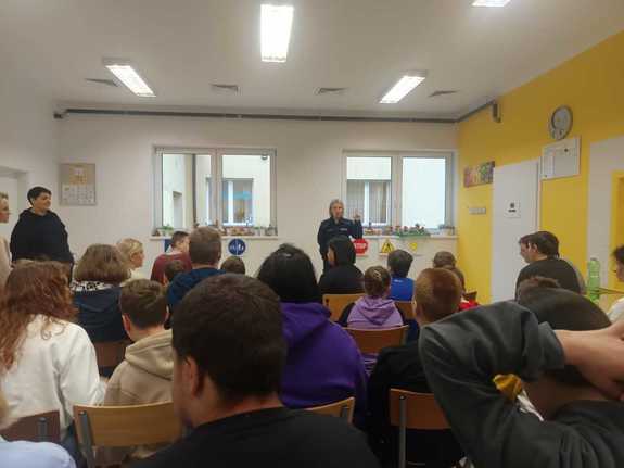 Policjantka i obok niej stojące znaki drogowe w sali na przeciw siedzącej młodzieży.