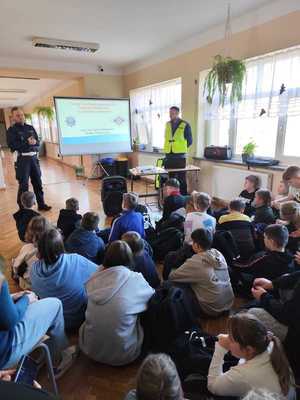 Dwóch policjantów przy tablicy na której wyświetlana jest prezentacja na przeciw siedzącej na sali młodzieży.