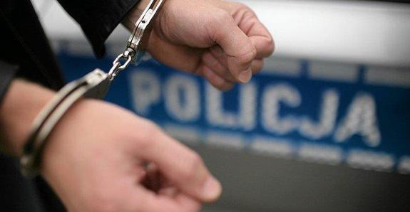 Na ręce założone policyjne kajdanki. W tle napis policja z fragmentem radiowozu.