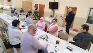 Policjant na sali stojąc przekazuje siedzącym przy stole seniorom informacje profilaktyczne.