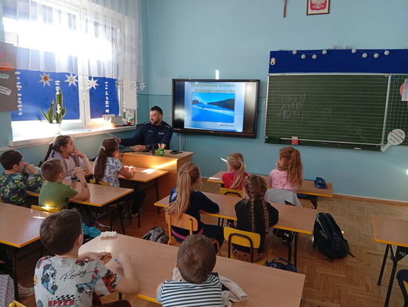 Policjant siedząc w klasie omawia dzieciom wyświetlaną na tablicy prezentację o bezpieczeństwie w zimie.
