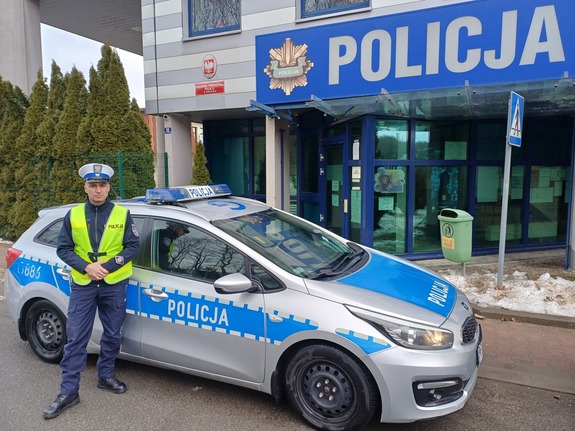 Policjant stojący przy radiowozie przed komendą Policji.
