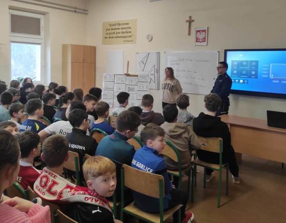 Policjant stoi w klasie przy tablicy  na prelekcji Kibicuj z klasą. Dzieci siedzą w ławkach.