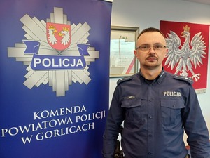 Policjant stoi przy logo gorlickiej Policji. Pod spodem napis Komenda Powiatowa Policji w Gorlicach.