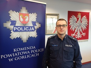 Policjant obok baneru z logo gorlickiej policji. Pod spodem napis Komenda Powiatowa Policji w Gorlicach.