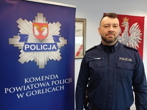 Policjant stoi przy banerze z gwiazdą policyjną i napisem Komenda Powiatowa Policji w Gorlicach.