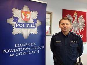 Policjant stoi obok baneru z gwiazdą policyjną i napisem Komenda Powiatowa Policji w Gorlicach.