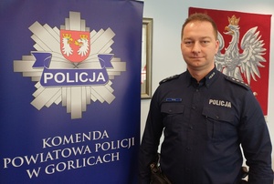 Policjant stoi obok baneru reklamowego z gwiazdą policji i napisem Komenda Powiatowa Policji w Gorlicach.
