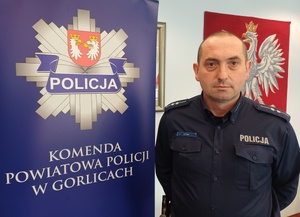 Policjant stoi obok baneru reklamowego z gwiazdą policji i napisem Komenda Powiatowa Policji w Gorlicach.