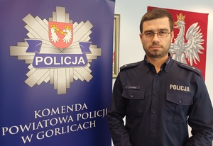 Policjant stoi obok baneru reklamowego z gwiazdą policji i napisem Komenda Powiatowa Policji w Gorlicach.