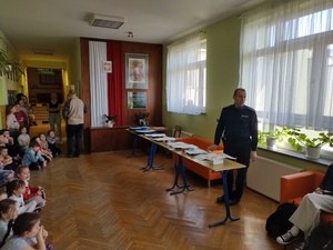 Policjant stojąc przy stole prowadzi prelekcję, dzieci siedzą wokół.