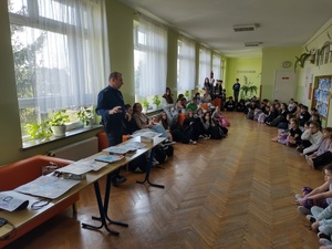 Policjant stojąc przy stole z nagrodami i wykonanymi pracami na konkurs prowadzi prelekcję.