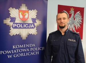 Policjant stoi obok baneru reklamowego za napisem komenda powiatowa policji w Gorlicach.