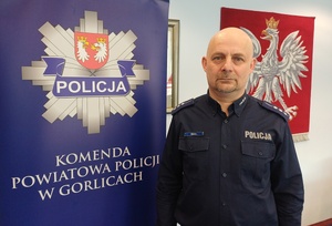 Policjant stoi obok baneru reklamowego z policyjna gwiazdą i napisem komenda powiatowa policji w Gorlicach.