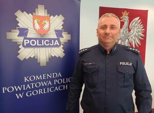 Policjant stoi obok baneru reklamowego z policyjna gwiazdą i napisem Komenda Powiatowa Policji w Gorlicach.