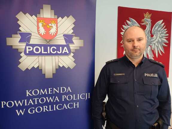 Policjant stoi przy banerze reklamowym z policyjna gwiazdą i napisem Komenda Powiatowa Policji w Gorlicach.