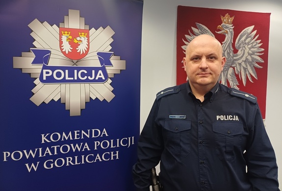 Policjant stoi obok baneru reklamowego z policyjną gwiazdą i napisem Komenda Powiatowa Policji w Gorlicach.