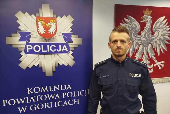 Policjant stoi obok baneru reklamowego z gwiazdą policyjną i napisem Komenda Powiatowa Policji w Gorlicach.
