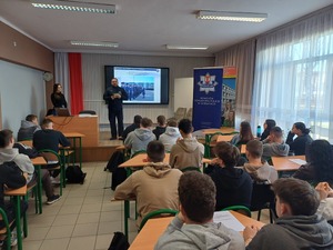 Uczniowie szkoły siedzą w ławkach, a policjant stojąc obok tablicy interaktywnej mówi do zebranych.