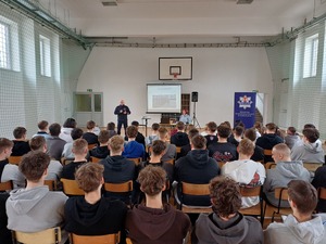 Policjant stoi obok stolika i prowadzi prelekcje do siedzących na sali uczniów.