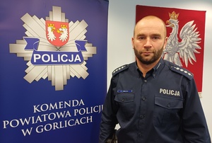 Policjant stoi obok baneru reklamowego z policyjną gwiazdą i napisem Komenda Powiatowa Policji w Gorlicach.