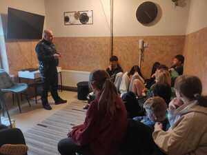 Policjant stoi w sali i prowadzi rozmowę z dziećmi siedzącymi na przeciw.